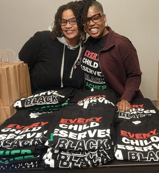 Two smiling individuals standing behind a table with stacks of t-shirts with the slogan "every child deserves a black teacher.