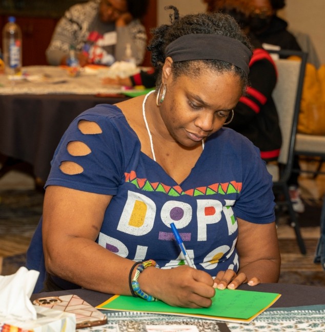 A woman in a blue t-shirt with the word "dope" written on it is focused on writing in a notebook at a table.