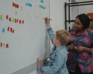 A woman observing a young boy as he writes on a whiteboard with colorful magnetic letters attached to it.