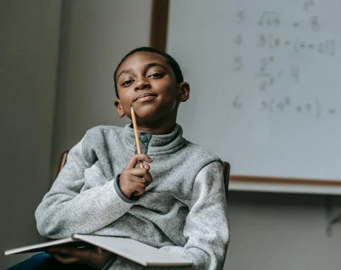 A thoughtful student holding a pencil and a notebook in a classroom with a whiteboard containing mathematical equations in the background.