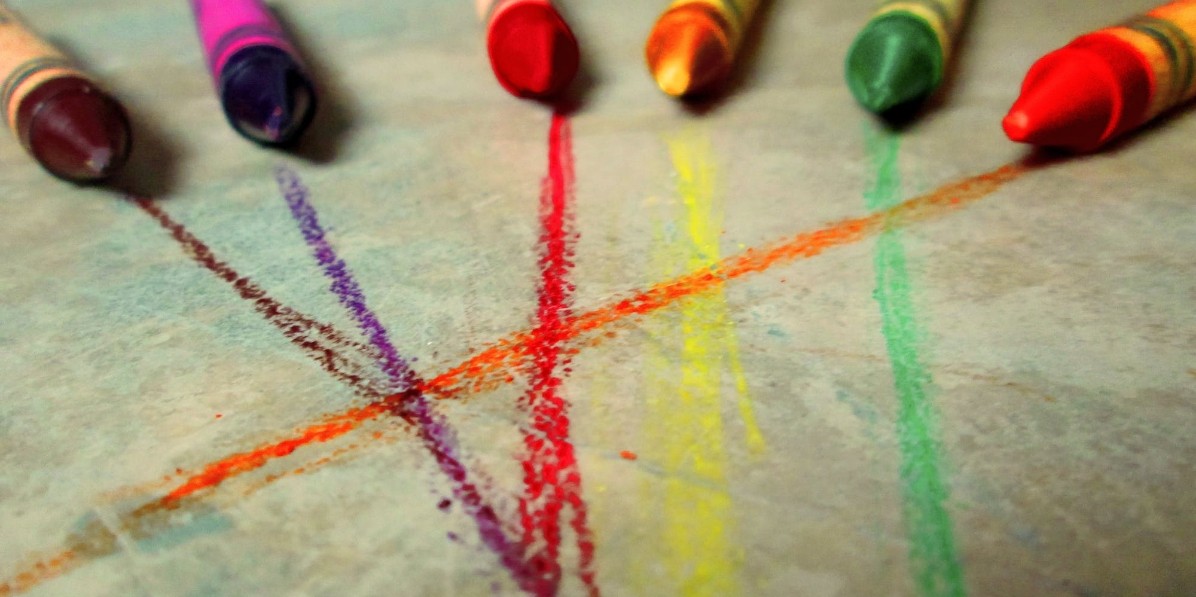 Colorful wax crayons arranged with their tips pointing towards the center of a playful tic-tac-toe game drawn on paper.