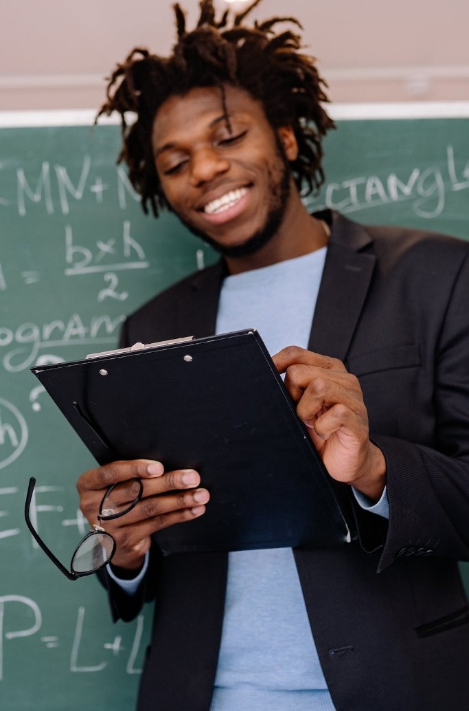 A smiling person holding a clipboard and glasses in front of a chalkboard with mathematical equations.