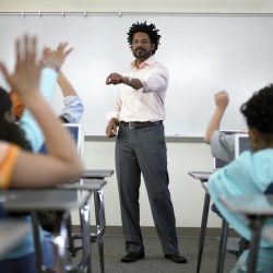 Male teacher standing before students (8-10) with hands raised